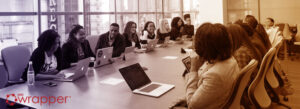 A large, diverse team of medical device professionals sitting at a long conference table with laptops, representing a coordinated effort for a remote QMS audit and virtual facility review.