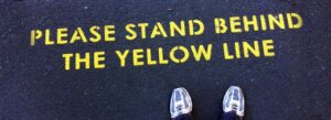 A yellow caution line on pavement with "PLEASE STAND BEHIND THE YELLOW LINE" text, serving as a metaphor for the risks of using unvalidated ISO 13485 template kits instead of a process-driven QMS solution.